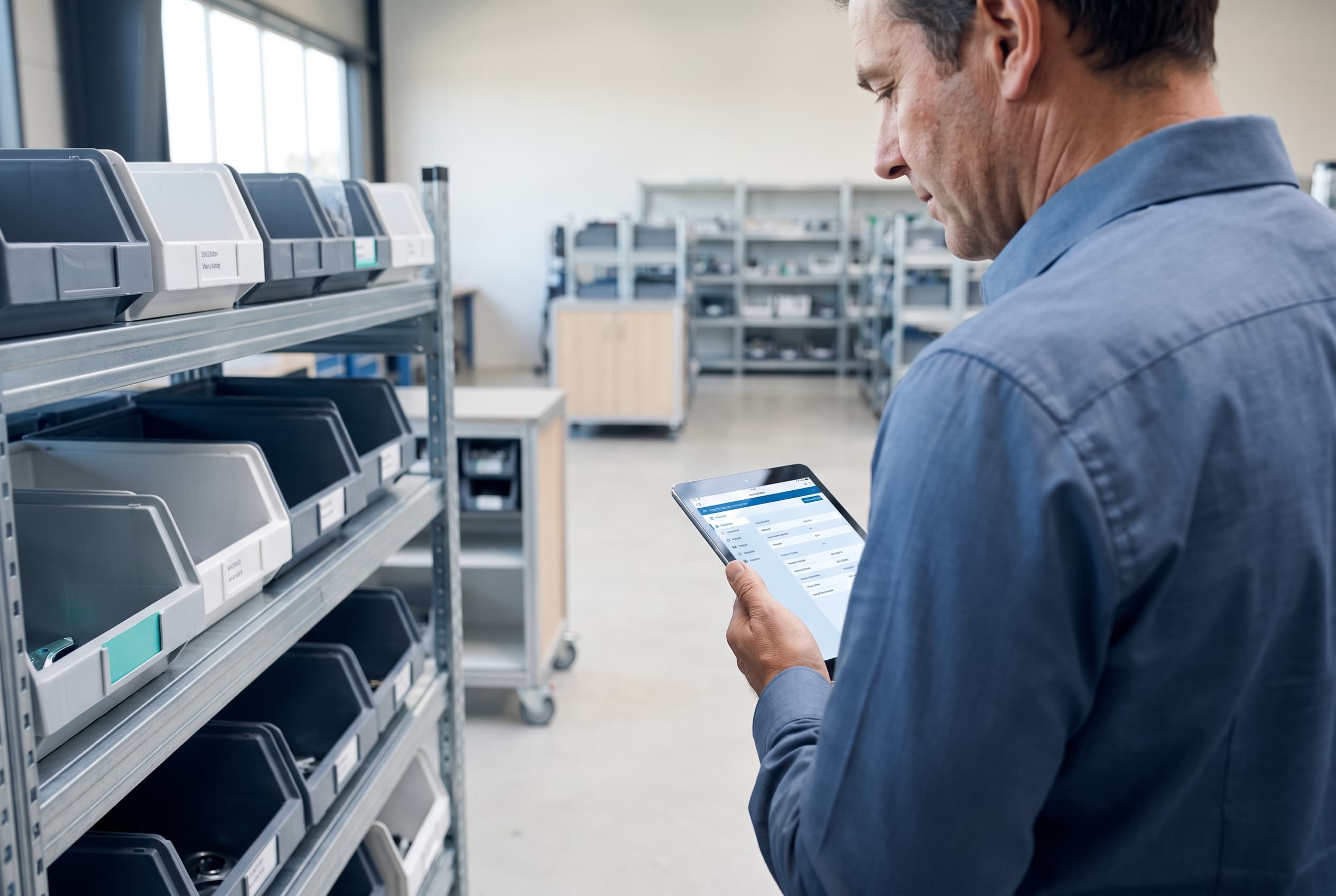 Un responsable des opérations consulte une affaire fabriquée à la commande sur une tablette, à côté d'étagères de pièces étiquetées dans un atelier