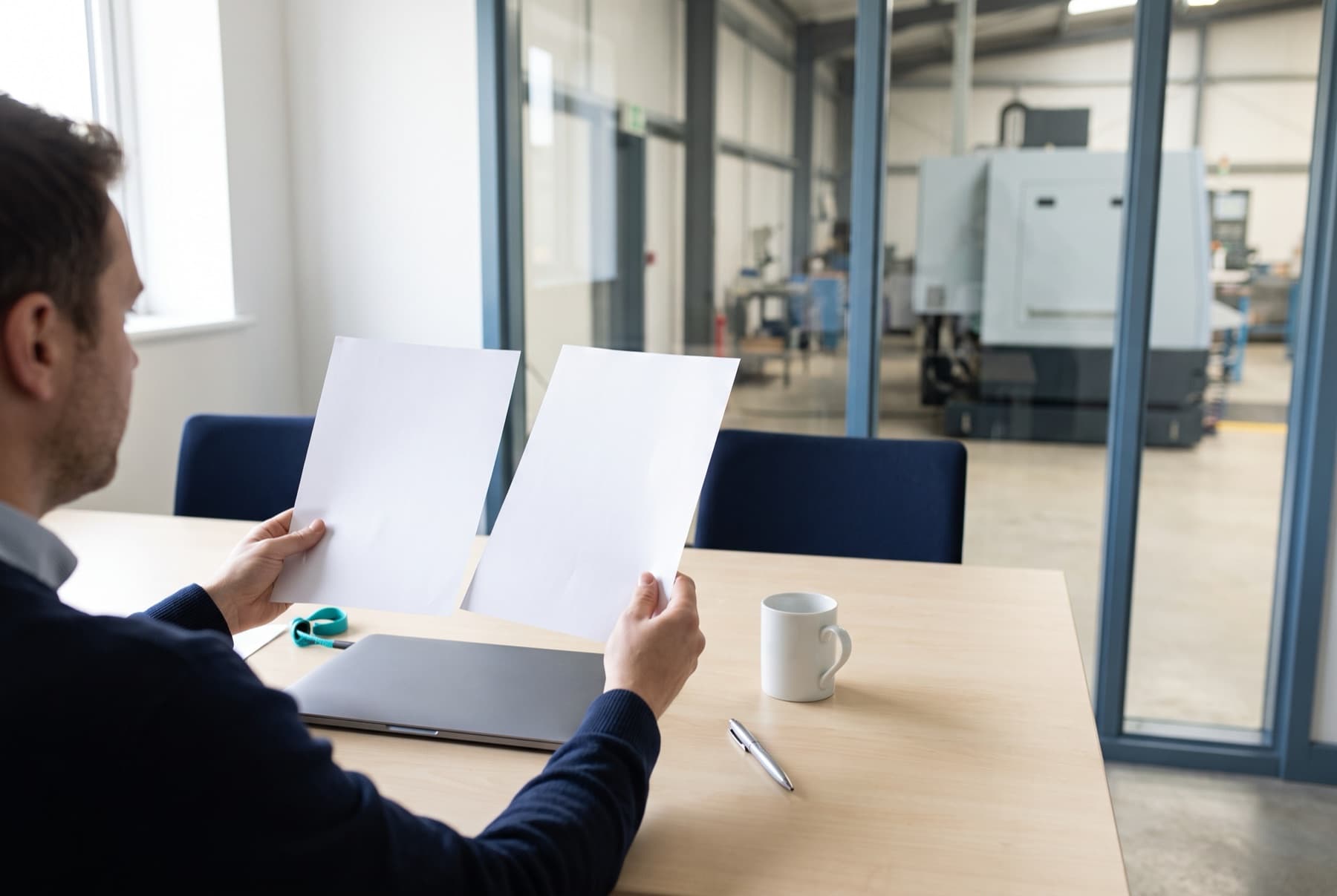 Responsable financier dans un bureau d'usine comparant deux documents côte à côte, l'atelier visible derrière la vitre